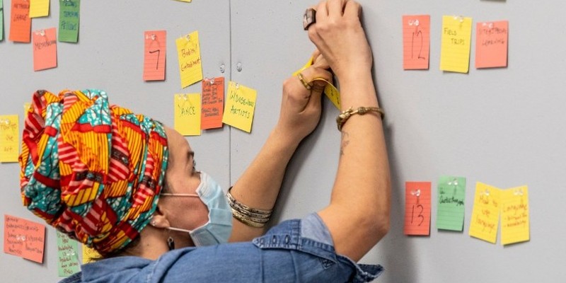 A woman in a brightly colored head wrap is writing on a post it note that is stuck to a gray wall. There are several other colorful post-it notes scattered around it.