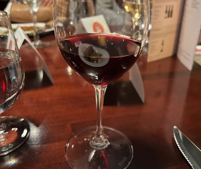 A glass with red wine sits on a dark wood table amongst other glasses and dishes.