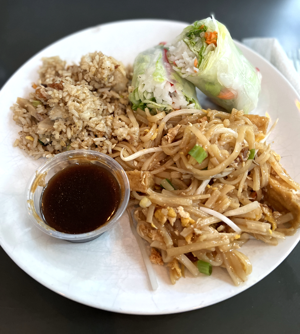 A round, white plate with portions of fried rice, Thai spring roll cut in half, and pad Thai. There is a small plastic container with a dipping sauce on the plate as well. Photo by Jessica Hammie. 