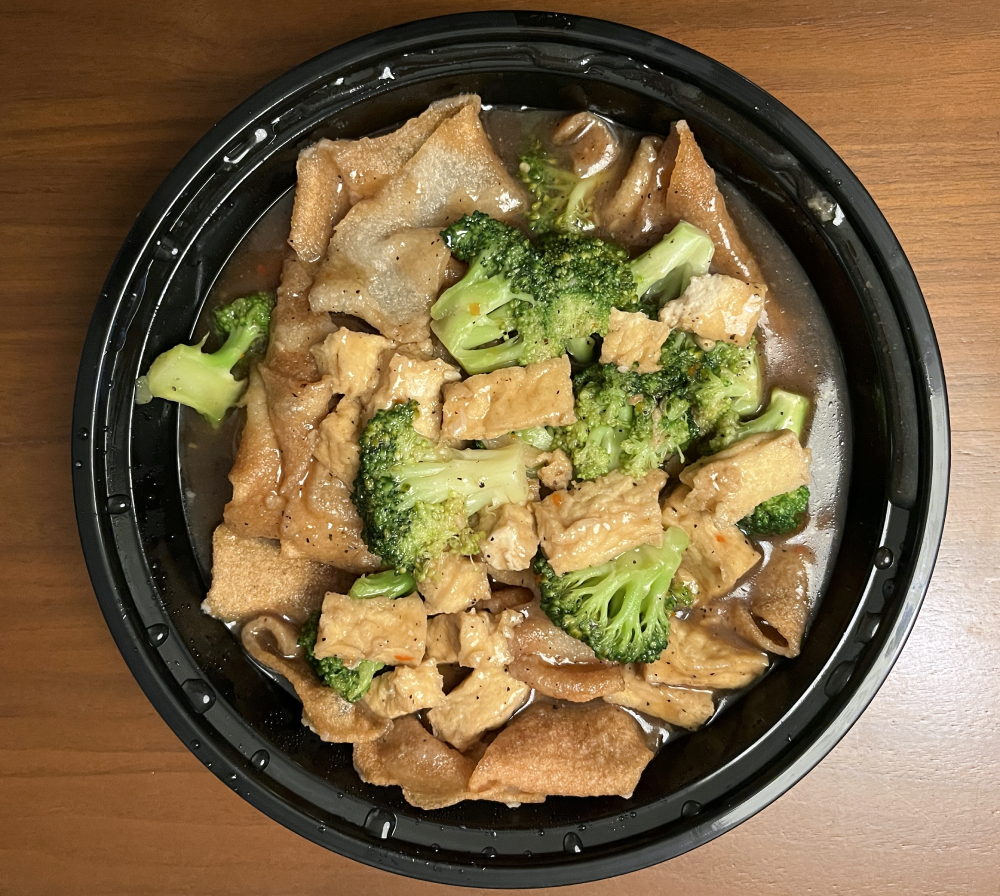 Pad lard nar in a round, black plastic take out container on a wood table. The dish includes wide noodles, tofu, and broccoli in a dark sauce. Photo by Jessica Hammie. 