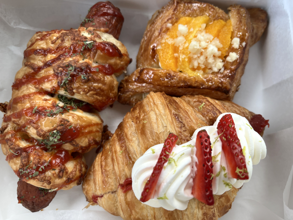 Clockwise, left to right: strawberry mascarpone croissant, an orange macadamia danish, and a hot dog croissant from BakeLab in Urbana. These three pastries are in a white takeout box. Photo by Jessica Hammie. 