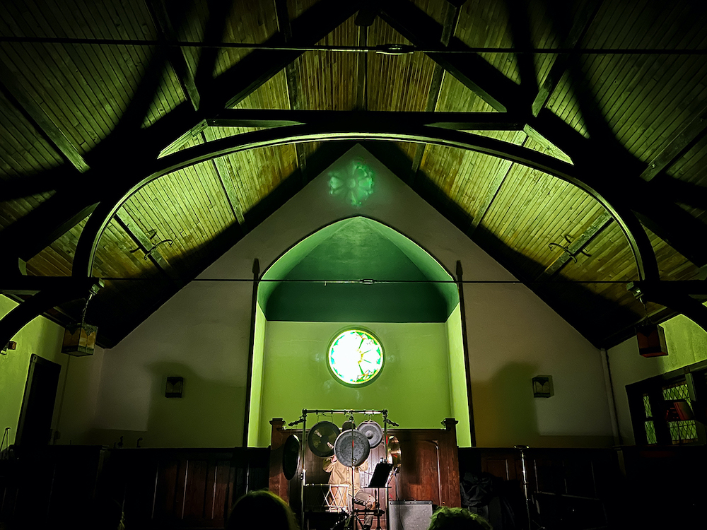 A view from the audience of warm light shifting between gold and green with a stained glass window below the rafters and above the Wisconsin based percussionist Jon Mueller behind his instruments - gongs, bells, drums- performing his recently released album The Future is Unlimited, Always on stage at The Channing Murray Foundation on Sunday, October 16th, 2022.