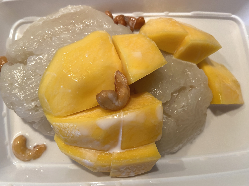 Mango sticky rice. Sticky rice is topped with large chunks of mango and some cashews in a white takeout container. The entire dish is drizzled with coconut milk. Photo by Jessica Hammie. 