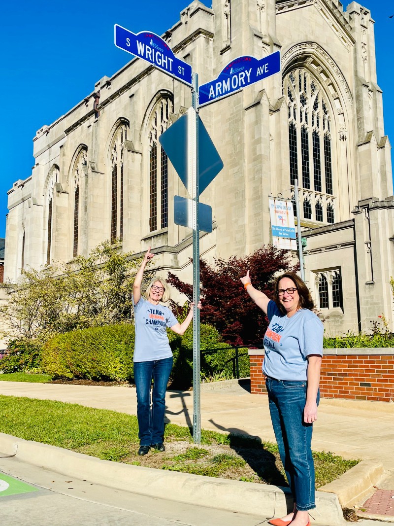 A woman with blonde hair and glasses and a woman with brown hair and glass are standing on either side of a street sign pointing up at it. There are two signs crossed, one that says Wright St and one that says Armory Ave. Photo from Visit Champaign County.