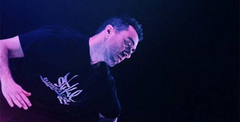 Close up photo of male dancer wearing a black t-shirt with white lettering mid-dance move left arm bending at the elbow, torso bending towards the left, against a dark blue black background.