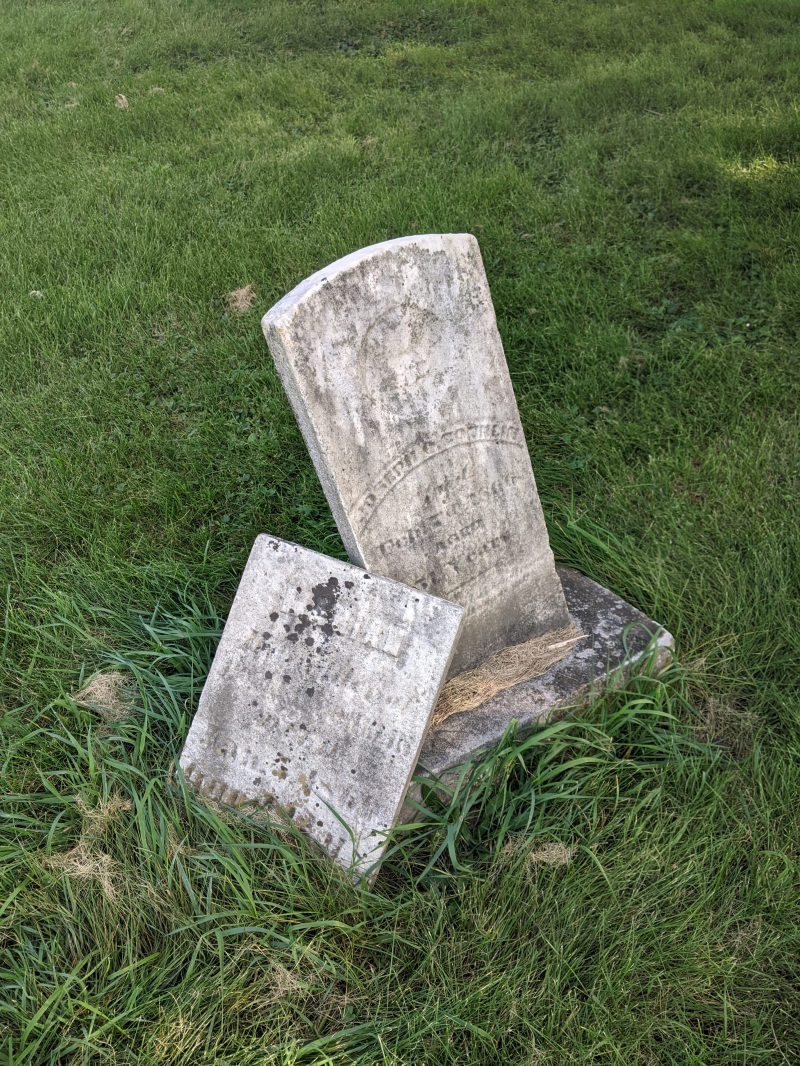 Two very old headstones that have partially sunk into the ground. They are leaning into each other and surrounded by green grass. Photo by Tom Ackerman.