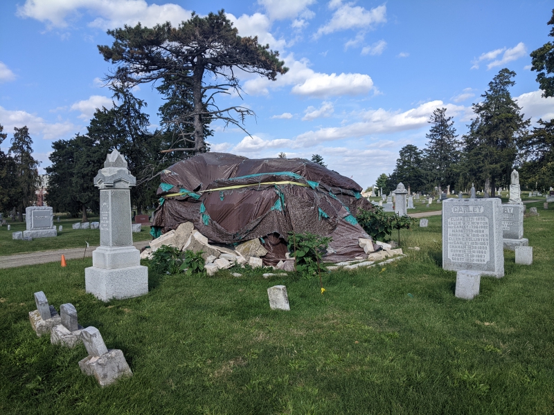 A crumbled stone mausoleum is covered in a large tarp and surrounded by other smaller headstones. Photo by Tom Ackerman. 
