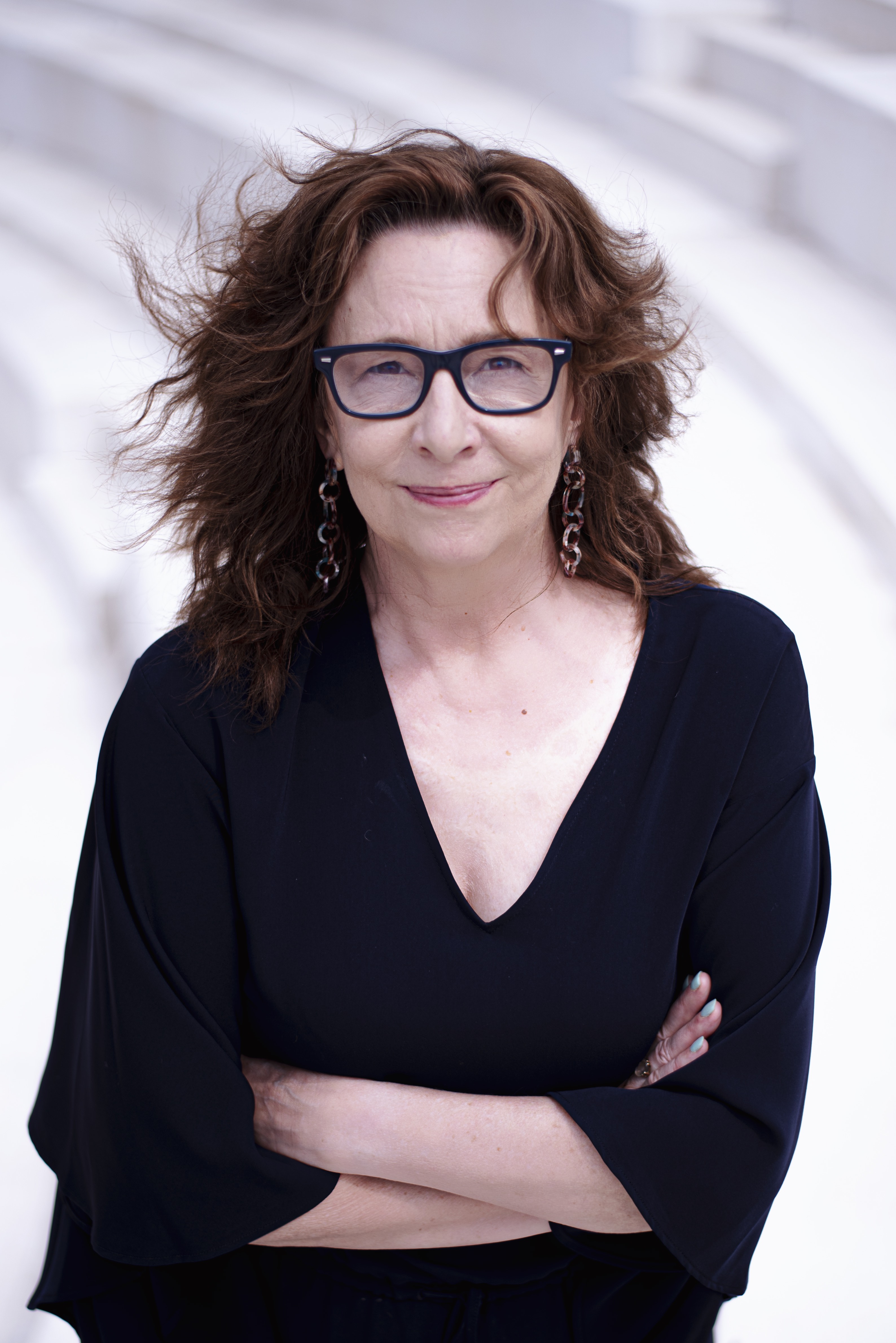 Portrait of Professor Sara J. Hook. She is a white woman with reddish-brown hair. She is wearing a black, v-neck sweater, dark framed glasses, and dangling statement earrings. Her arms are crossed in front of her body and she looks at the camera and gives a small smile. Photo by Elliot Emadian. 