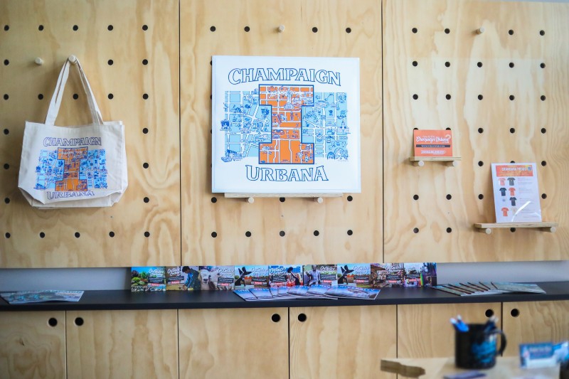 A light wood display wall with a tote bag and sign that have a map of Champaign-Urbana with an orange block I over it. Photo from Visit Champaign County.