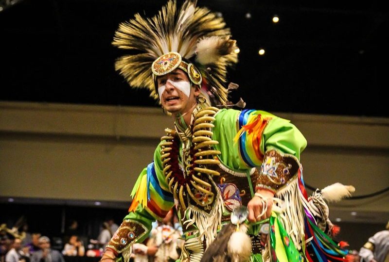 A man in Native American regalia, including a large feathered headpiece and brightly colored robes, is dancing.