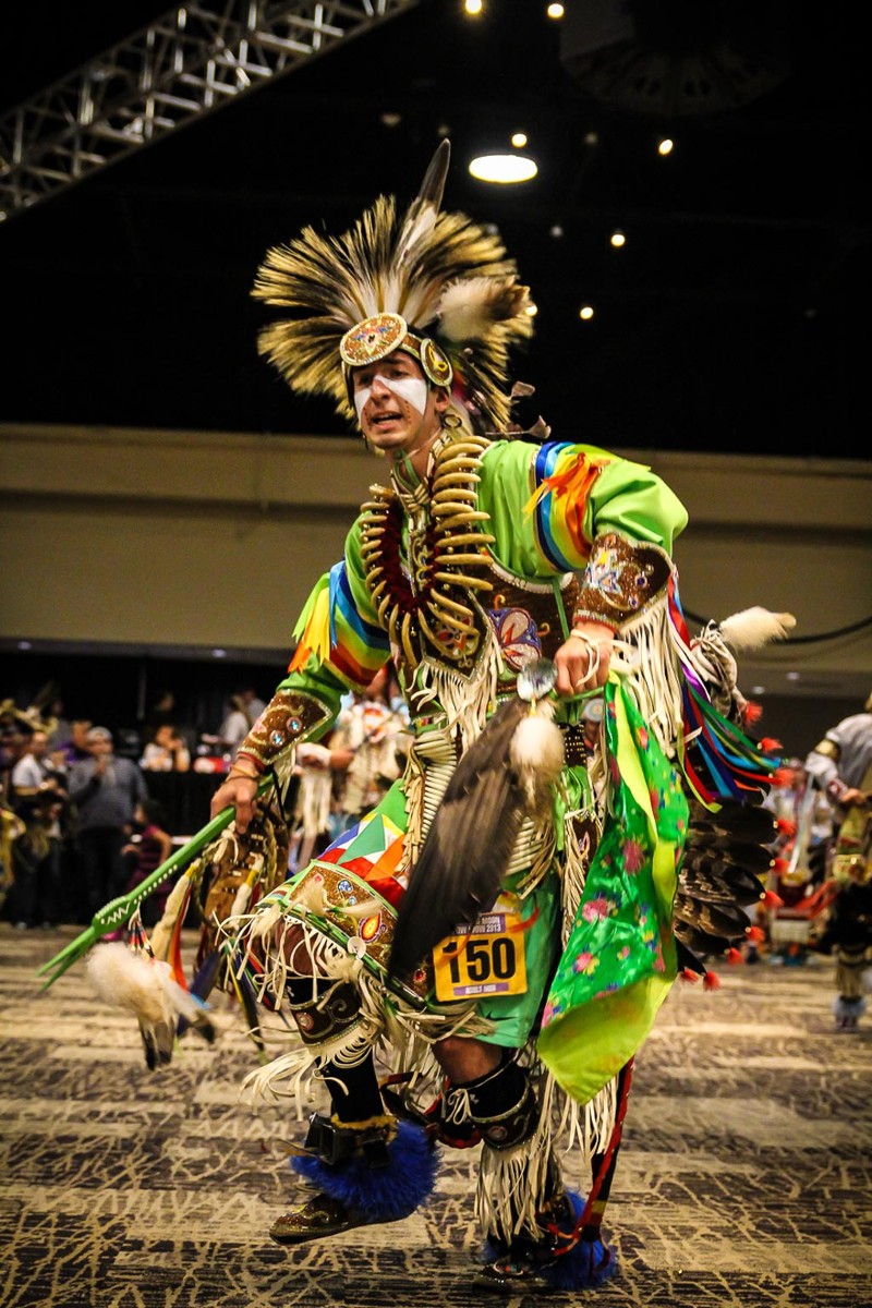 A man is dressed in full Native American pow-wow regalia, dancing in a large room with people watching. Photo from Spurlock Museum Facebook page.