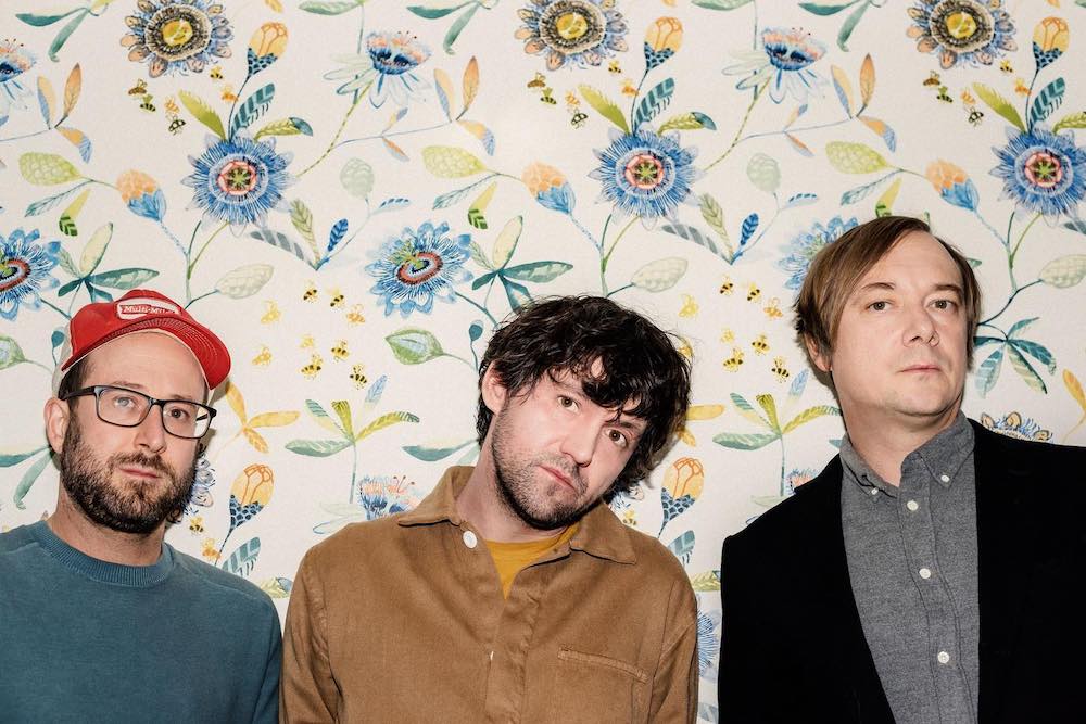 Portrait of the three members of bright eyes looking into the camera. They stand in front of a wall with yellow and blue floral decorations. 
