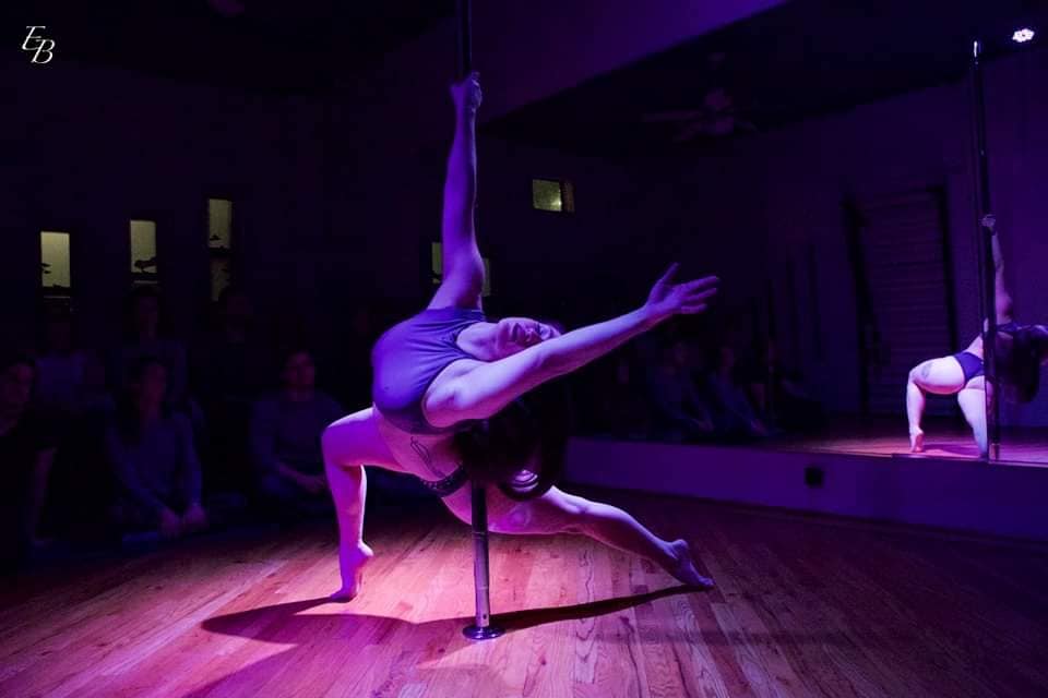 A woman is dancing around a pole; her upper body is extended toward the camera, and her lower body is wrapped behind the pole. Her left arm is extended, and her right arm is gripping the pole. Photo from Defy Gravityâ€™s Facebook page.