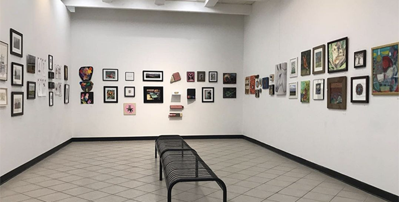 Photo of three white walls in an art gallery filled with art work of different sizes and shapes. A Black wire bench stands in the middle of the room.