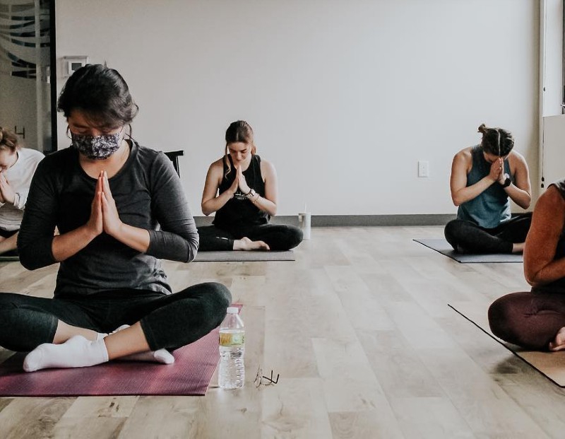 There are four women gathered in a space with wood floors. They are sitting on yoga mats, with hands in prayer position. Photo from Hatha Yoga and Fitness Facebook page.