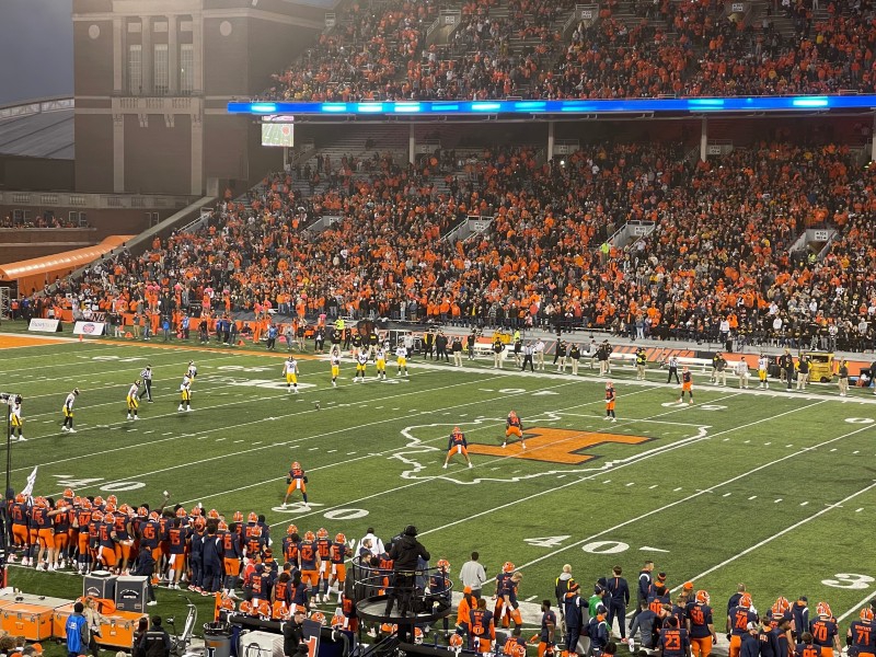 The field at Memorial Stadium during a football game. Players are line up on either side of the field, and the stands on either side are filled with people, many of them wearing orange. Photo by Julie McClure.