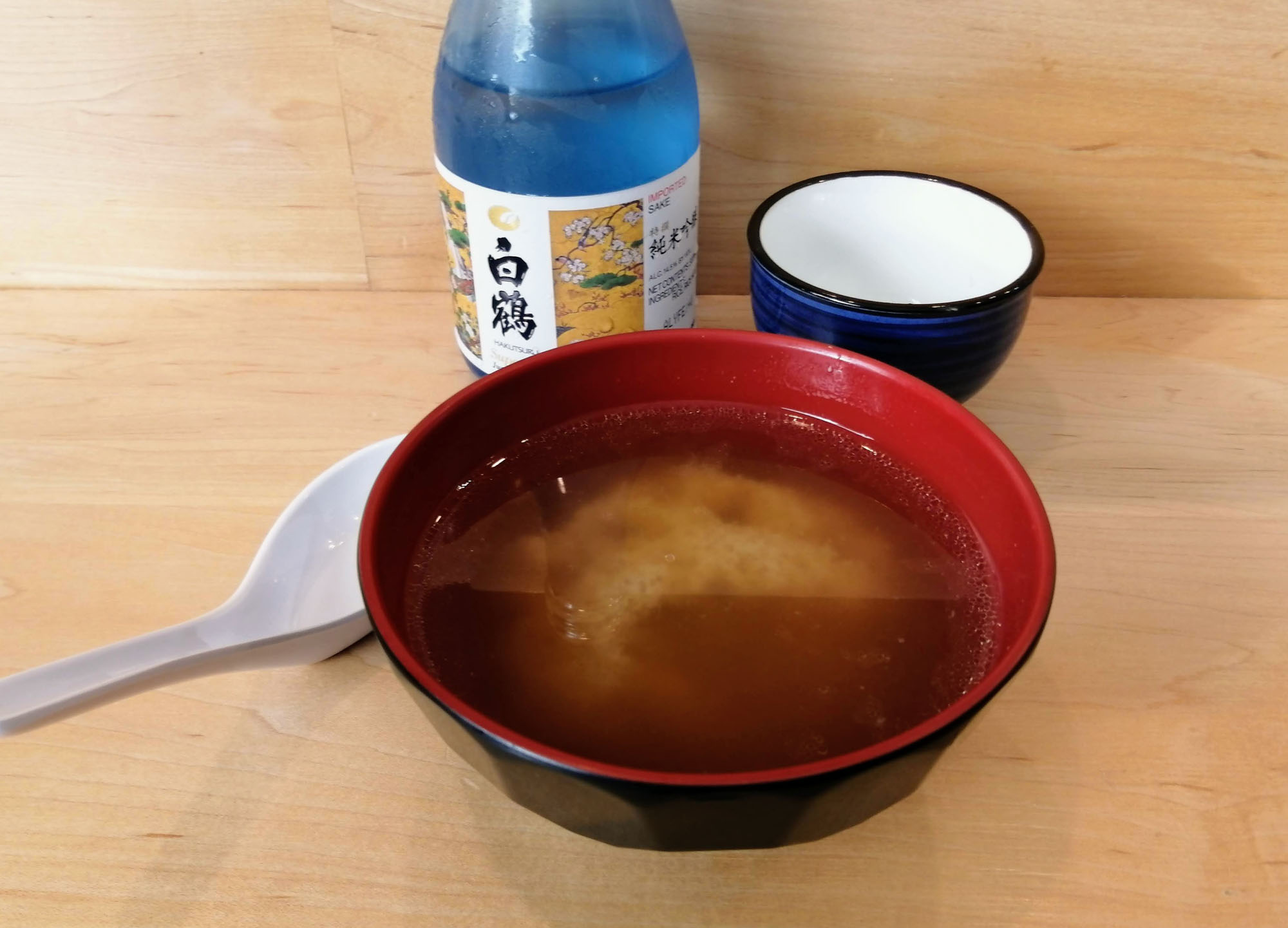 A close-up of a bowl of miso soup with a bottle of sake and a cup visible in the background. Photo by Paul Young.