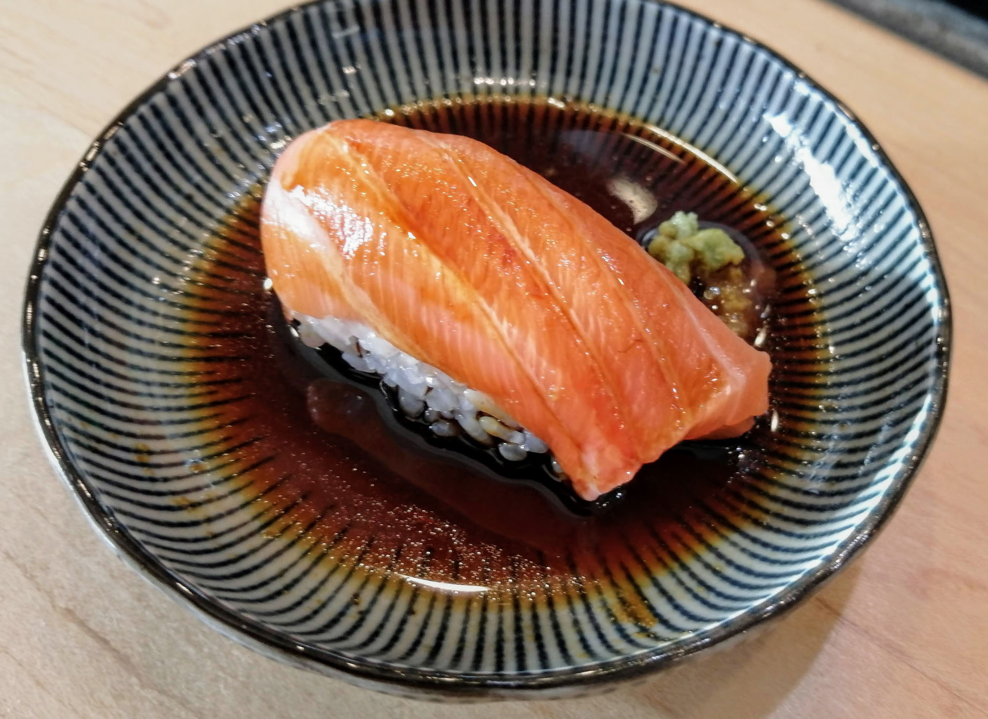 A close-up of a piece of nigiri sushi made with a slice of raw salmon sitting on a serving plate with soy sauce and wasabi. Photo by Paul Young.
