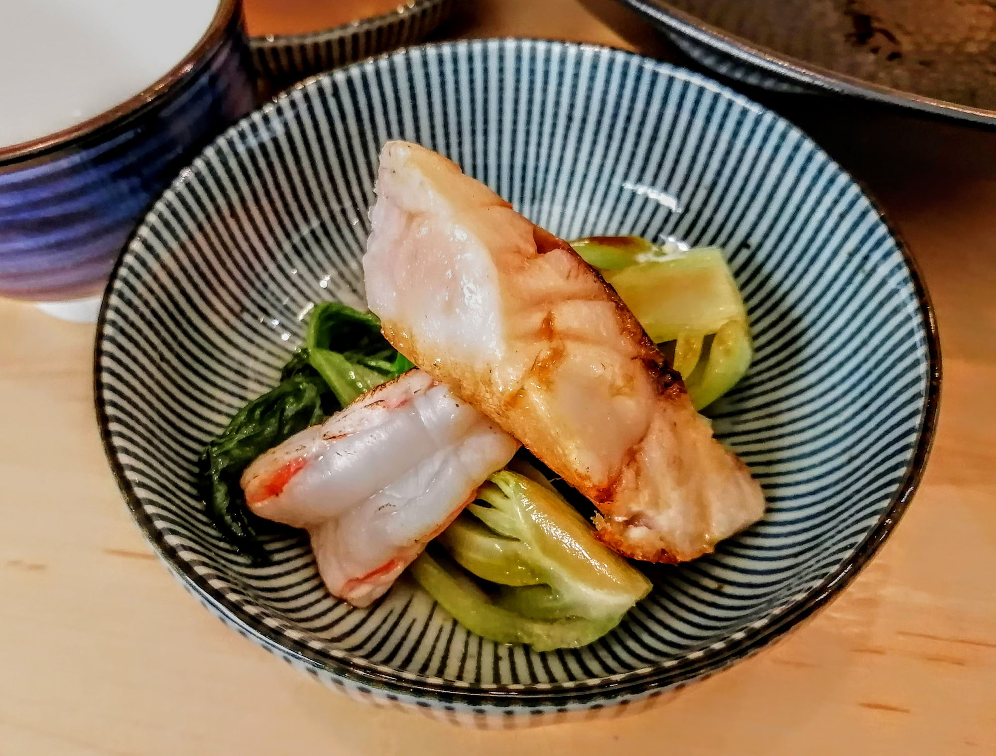 A close-up of seared fish, shrimp, and bok choy nestled inside a striped bowl. Photo by Paul Young.