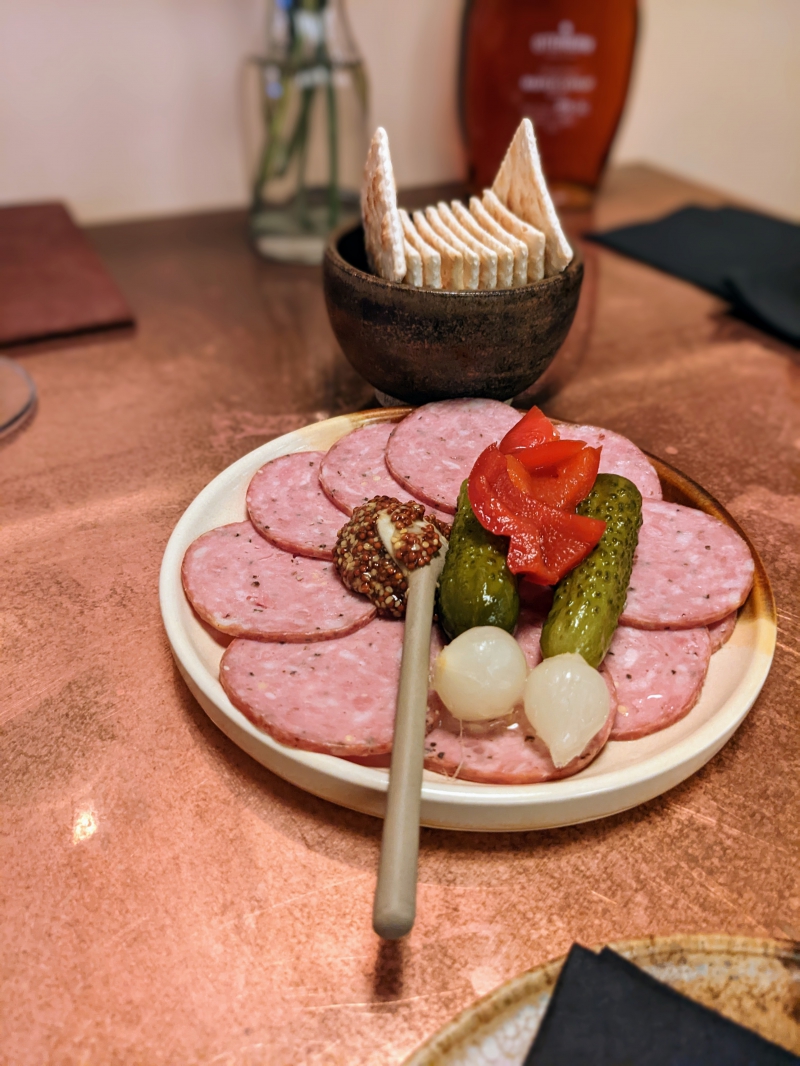 On a white handmade plate, summer sausage is sliced and arranged beautifully beside grainy mustard and pickles. Photo by Cailtlin Aylmer.