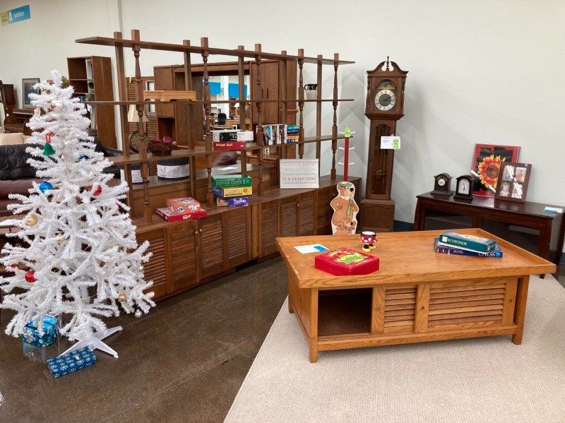 A display set up to look like a living room, with a rug, wooden coffee table with games on it, a white Christmas tree, a grandfather clock, and large wooden shelving unit. Photo from Champaign County Restore Facebook page.