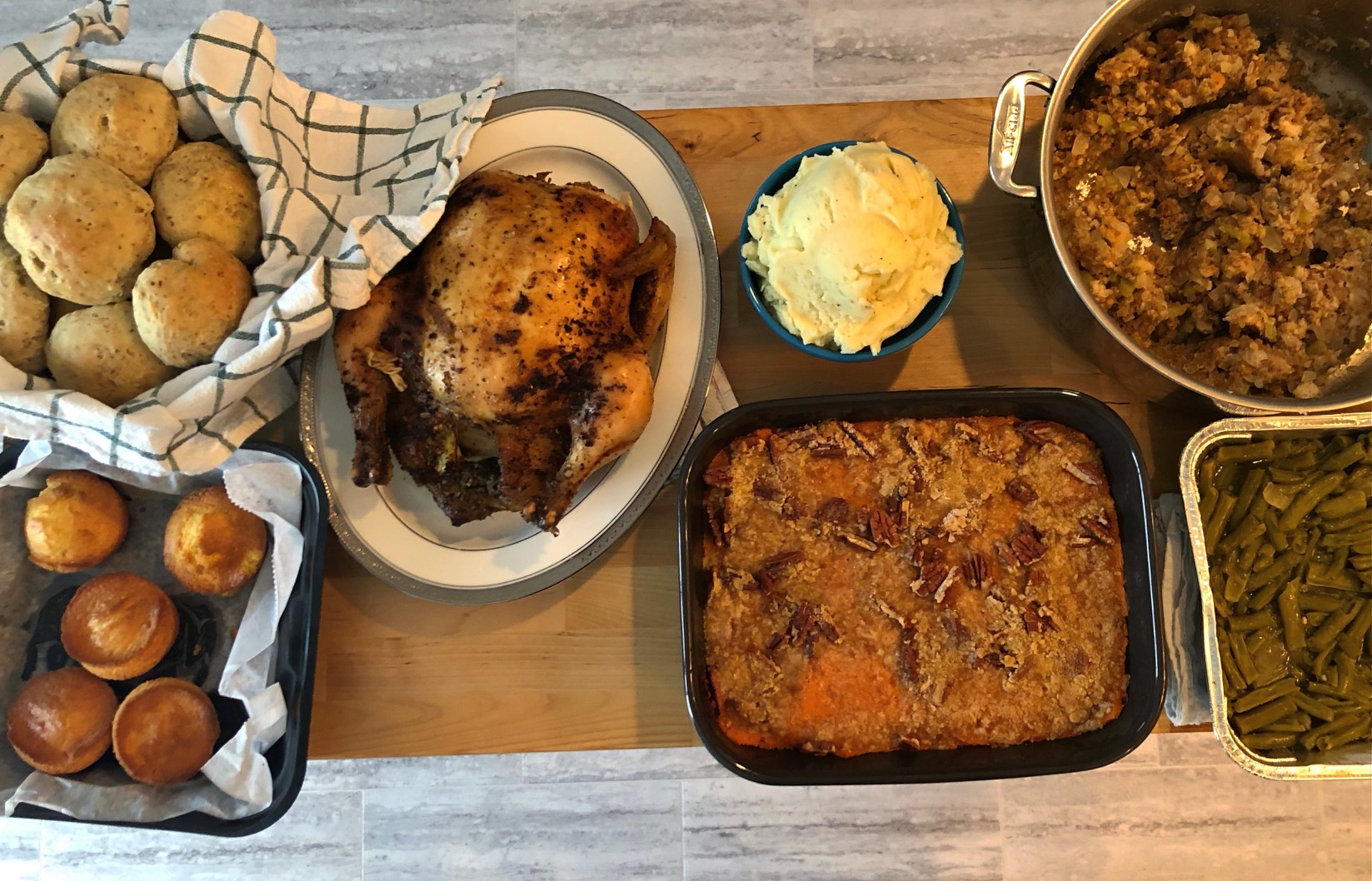 A Thanksgiving dinner table as seen from above. On the wood table are two different types of rolls, a roasted turkey, mashed potatoes, sweet potato casserole, stuffing, and green beans. Photo by Alyssa Buckley.