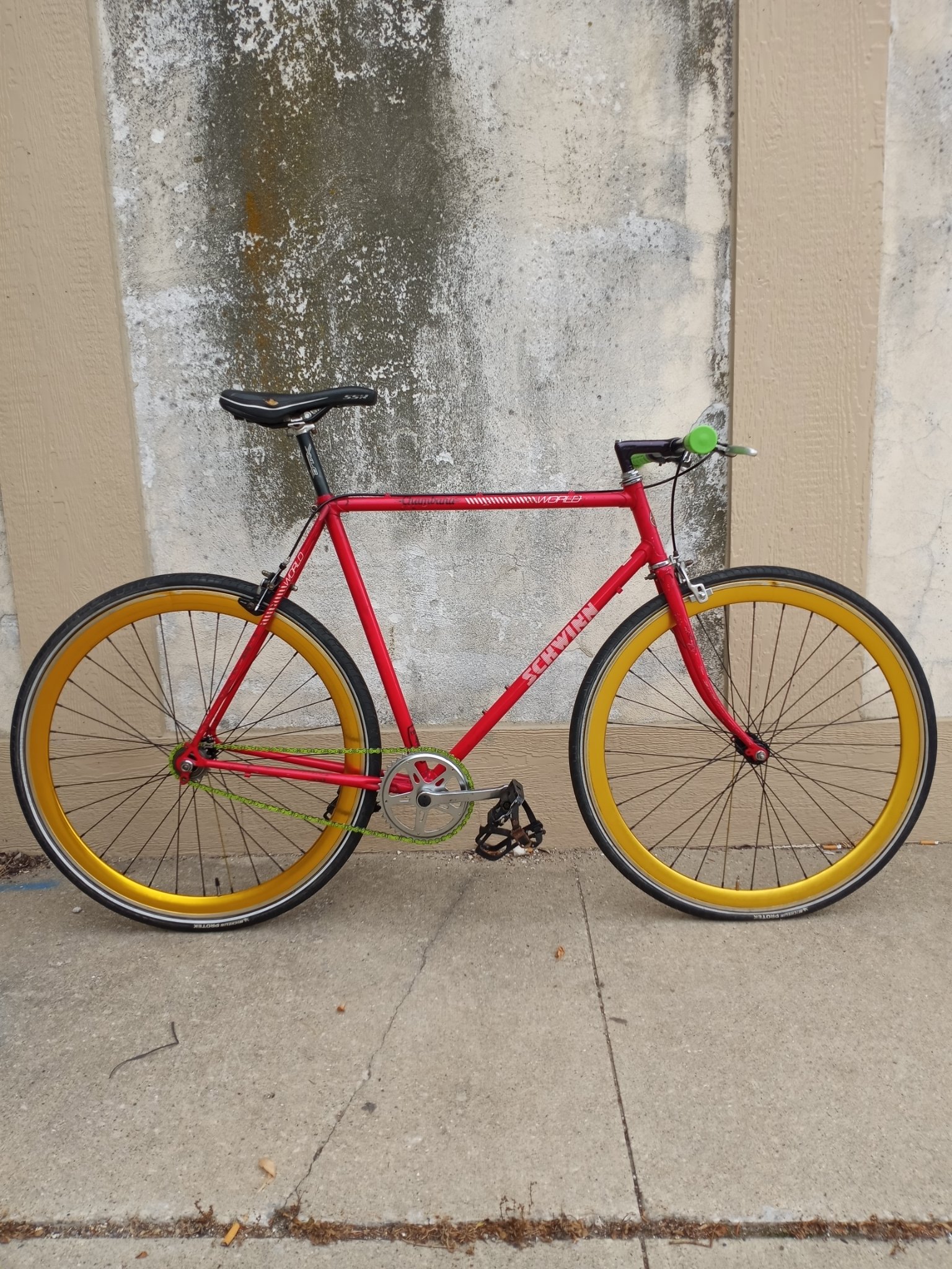A red bicycle with yellow wheels and black tires is leaning on a pale concrete wall, on a concrete sidewalk.