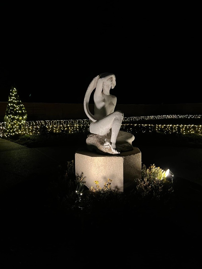 A stone figure sitting on a stone pedestal is encircled by white lights and evergreen trees with white lights. It is pitch dark. Photo by Julie McClure.