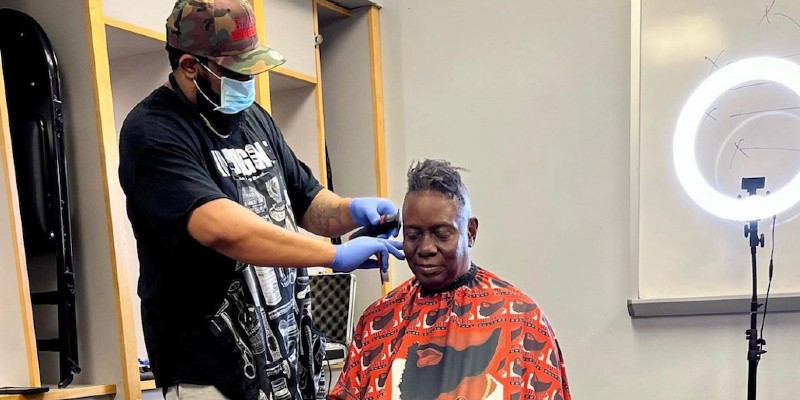 A Black man is sitting in a barber's chair, wearing a cape, while another Black man in a black t-shirt and camouflage ball cap and face mask is cutting his hair. Photo from Instagram.