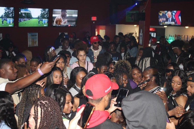 A large group of people are crowded on a dance floor in a bar. There are televisions lining the walls near the ceiling. Photo by Denzel McCauley.