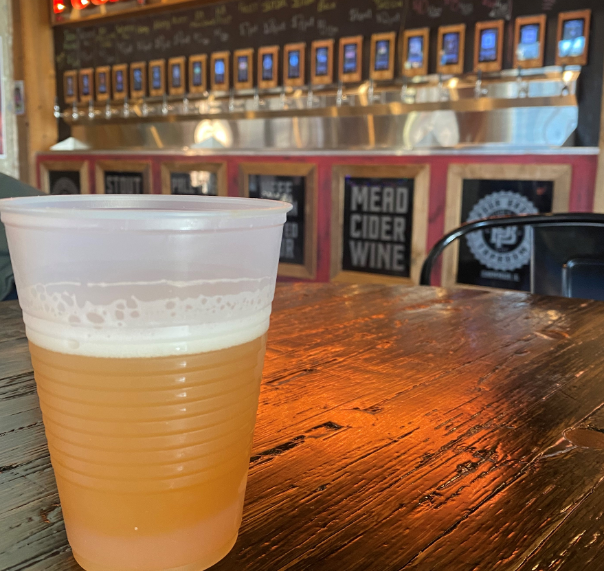 Blue Hours IPA. The light amber beer has a white head is in an opaque plastic cup on a wood table. In the background, a row of labeled taps dispensing different beverages is visible. Photo by Destini Merris.
