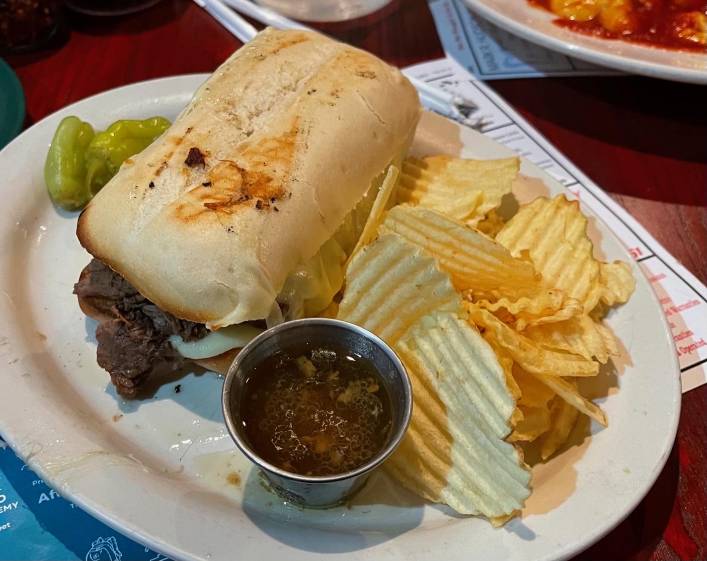 On a white plate, there is an Italian beef sandwich with ridged chips and a cup of au jus. Photo by Stephanie Wheatley.