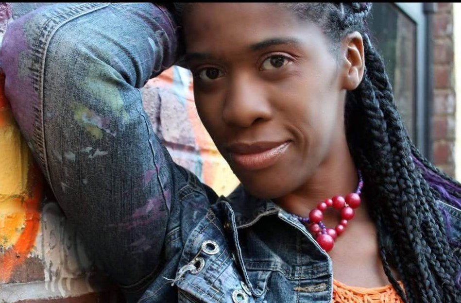 Ja Nelle Davenport-Pleasure portrait photo. She is a Black woman, with long braids. She is wearing a jean jacket, orange shirt, and red beaded necklace. Her gaze is looking at the camera with her arm up to her face. Behind her is a brick wall.