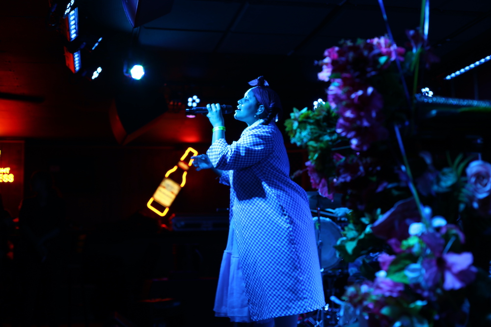 View from the side of a stage: A woman in a long checked coat is singing into a microphone. She is bathed in blue light. There is a neon beer sign in the background, and flower display in the foreground.