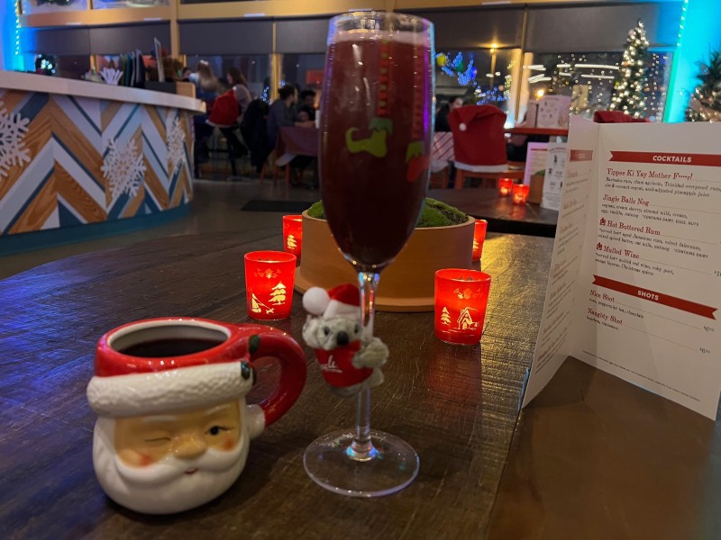 Two drinks sit on a low wooden table. One has a dark liquid in a ceramic Santa head mug. The other has a reddish-purple liquid in a clear champagne flute. There's a tiny stuffed koala wrapped around the stem. Photo by Julie McClure.