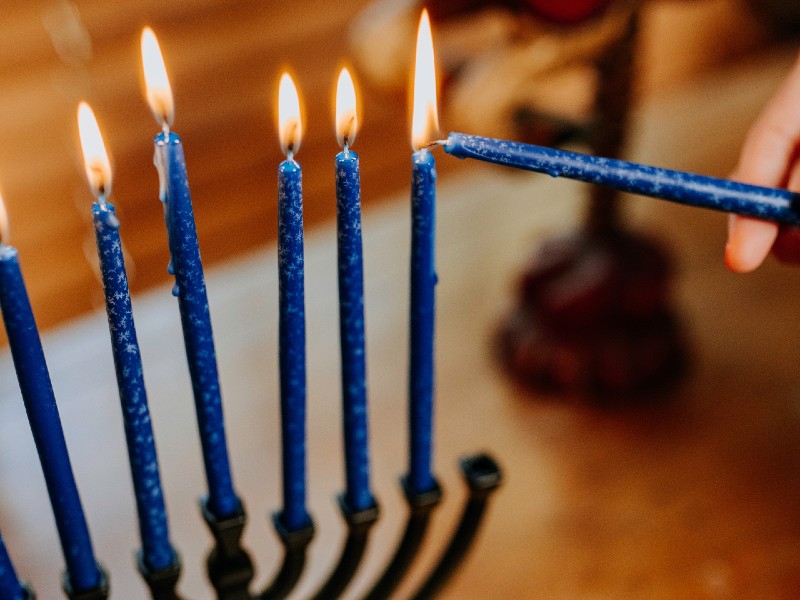 Close up of a portion of a menorah with thin blue candles that are lit. There is a hand holding another candle, and lighting it from another. Photo from pexels.com.