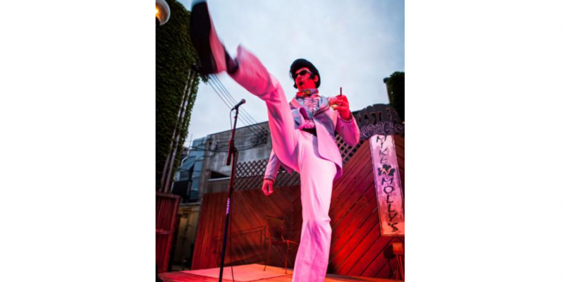 Aaron davidson on stage in a white suit, dressed as Elvis, kicking his leg out towards the audience. There is a microphone in front of him and a wooden panel behind him. Photo by Troy Stanger.