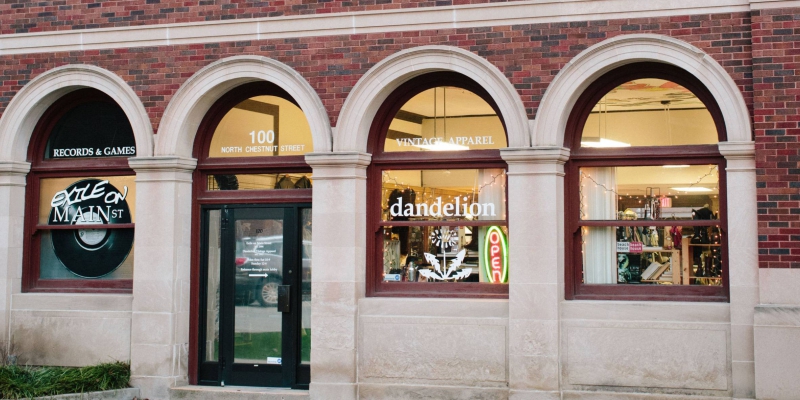  A storefront with three arched windows and an arched glass doorway and a red brick facade. The far left window has the words â€œExile on Mainâ€ in white lettering. A window to the right of the door says â€œdandelionâ€ in white lettering, with a white flower decal below it. There is a vertical neon â€œOpenâ€ sign. Photo by Anna Longworth.