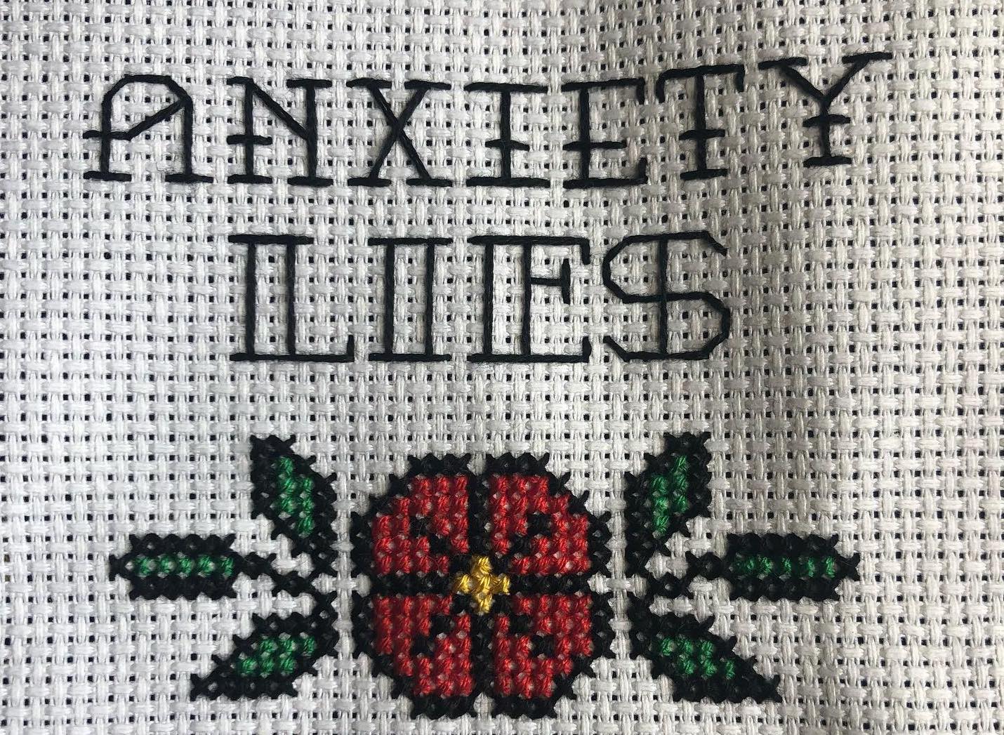 Cross stitch that reads "anxiety lies" with a red flower underneath. The background is white, the text is black.