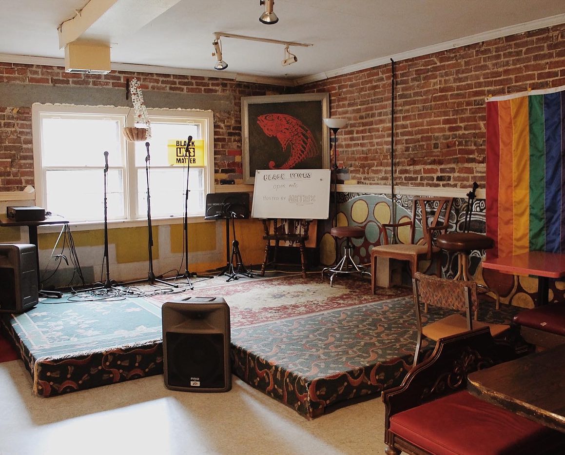 A view of the performance stage and walls behind it at The Red Herring restaurant. A stage covered in rugs is in the corner of the room. Mismatched chairs are on the right side. 