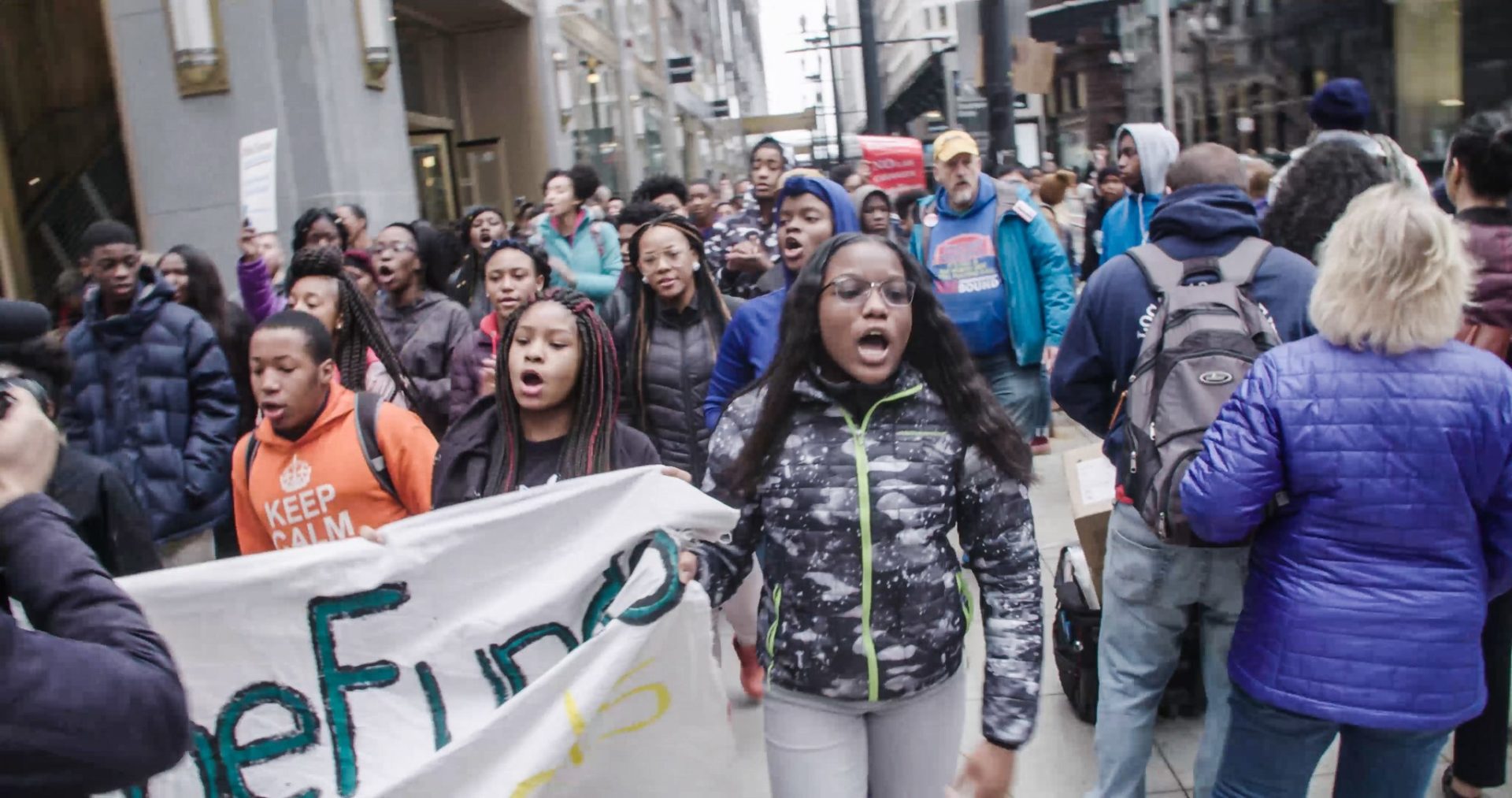 A crowd of mostly Black young people are marching down a city sidewalk with banners. Most of their mouths are open as if chanting.