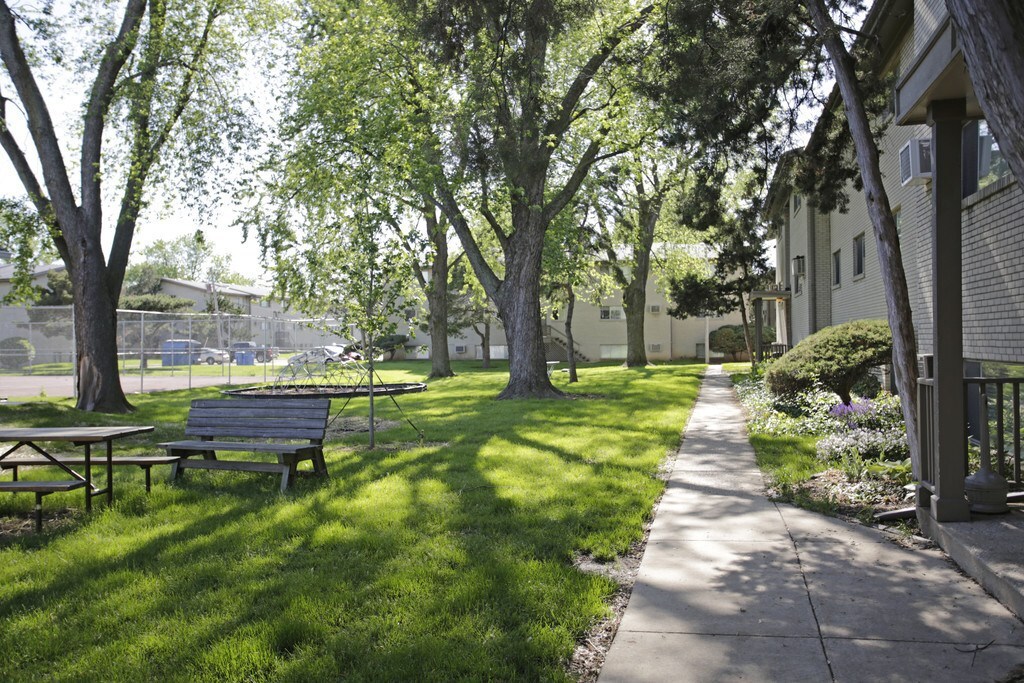 Outside photo of the Champaign Park Apartments. Green grass is on the left, there is a grey sidewalk in the middle, and apartment buildings to the right. There are picnic tables in the grass and a tennis courts in the background.