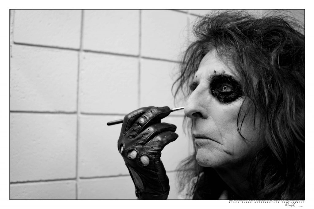 Black and white photo of musician Alice Cooper in profile, applying his signature eye makeup look.