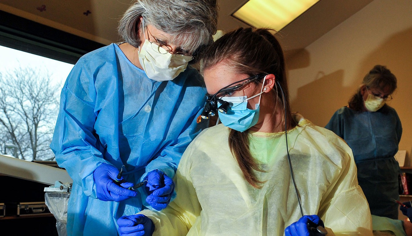 A woman with gray hair in blue scrubs, a mask, and blue rubber gloves is leaning in to show something to a younger woman with long brown hair. The young woman is wearing a yellow paper gown, a mask, and blue rubber gloves.