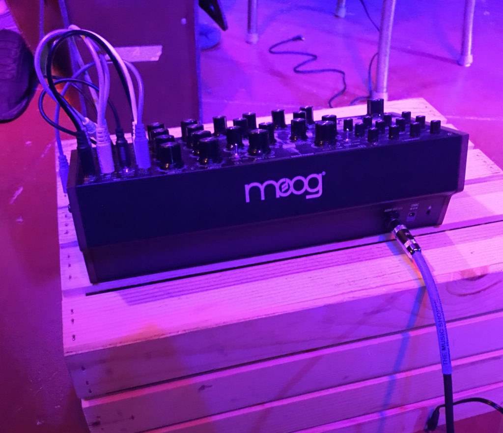 A Moog synthesizer is on a wood crate. There are several wires attached to the top, and cords from the back. It seems to be that the crate is on a stage, and the lighting is purple and pink. 