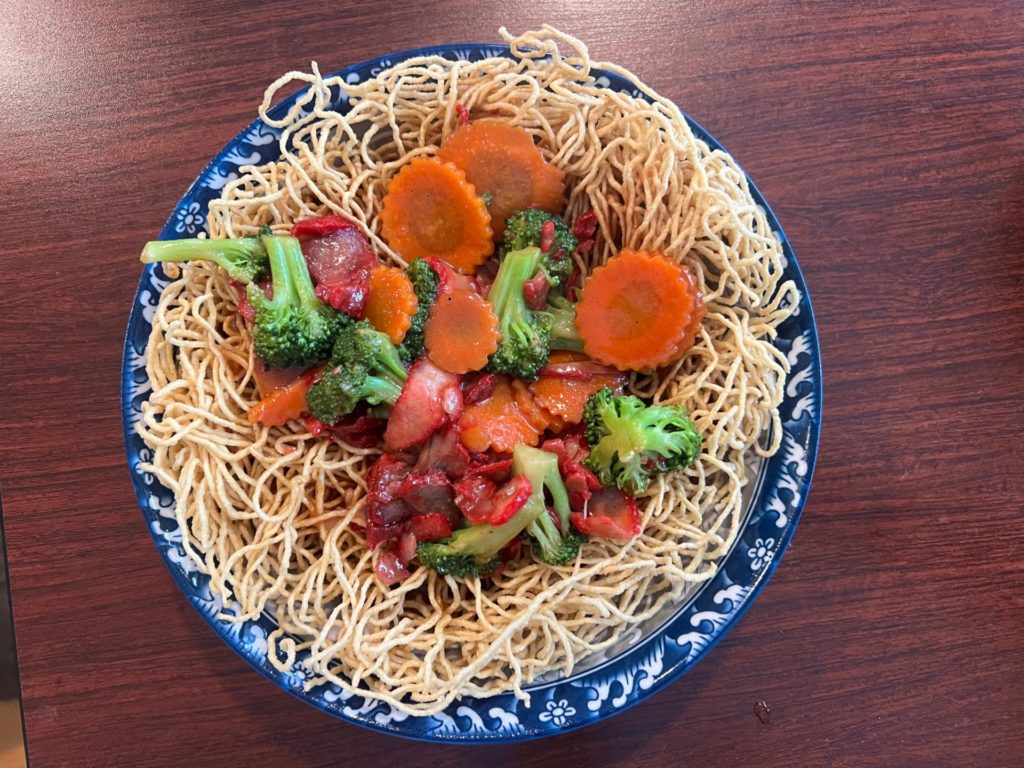 On a brown table, there is a blue and white bowl with a pretty rim holding mee korb from Lanxang in Champaign, Illinois. The mee korb has a nest of fried egg noodles with the center filled with pork and veggies. Photo by Alyssa Buckley.