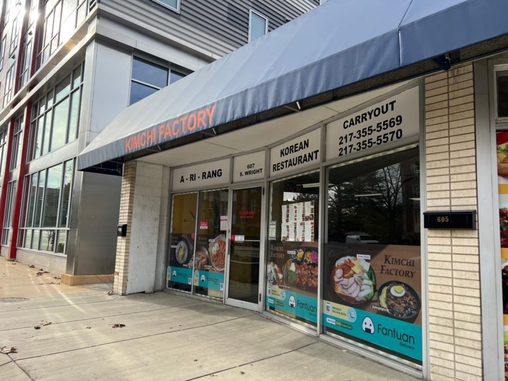 On Wright Street in Champaign, Illinois, the exterior of Kimchi Factory, previously A-Ri-Rang has a blue awning and large windows. The windows have carryout phone numbers, ads for delivery, and photos of Korean food. Photo by Alyssa Buckley.