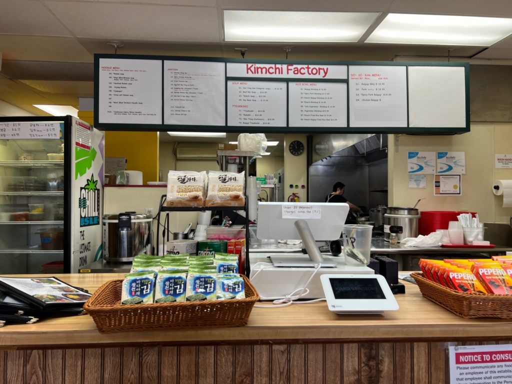 Inside Kimchi Factory in Champaign, Illinois, there is a counter with Korean snacks in wicker baskets. Behind the counter there is a white menu with black and red ink. The kitchen is open and the chef cooks over a stainless steel stove. Photo by Alyssa Buckley.