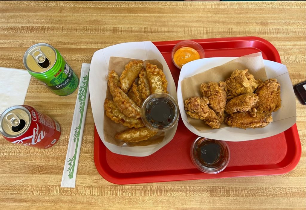 At Kimchi Factory, there are two appetizers in paper baskets lined with brown parchment paper. On the left is fried dumplings and on the right fried chicken wings. There are three small sauces: two dark brown and one orange in small plastic cups. All the food is on a bright red tray atop a wooden table. Also on the table are two cans: Coke and green tea plus a paper-wrapped pair of chopsticks. Photo by Alyssa Buckley.