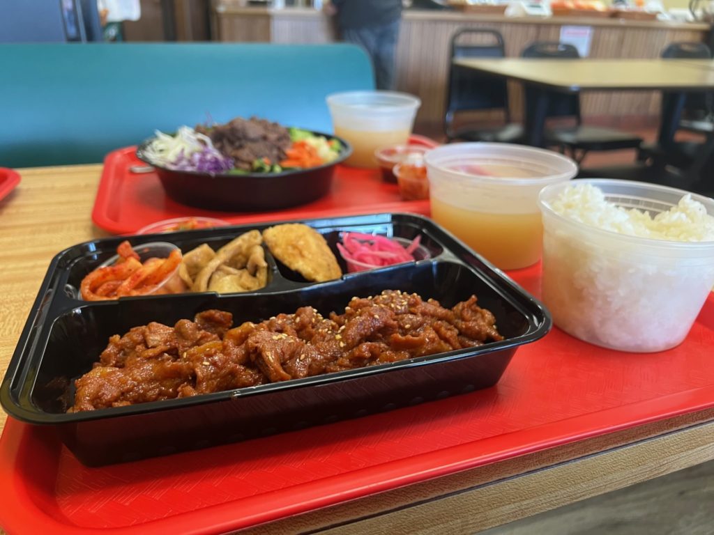 On a red tray, there is a divided black plastic container with spicy pork and a variety of banchan. Beside that there is a clear plastic cup of white rice and a clear plastic cup of brothy soup. Photo by Alyssa Buckley.